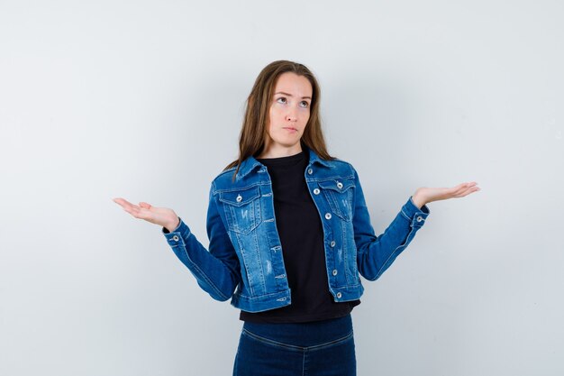 Jonge dame in shirt, jas die een weegschaalgebaar maakt en peinzend kijkt, vooraanzicht.