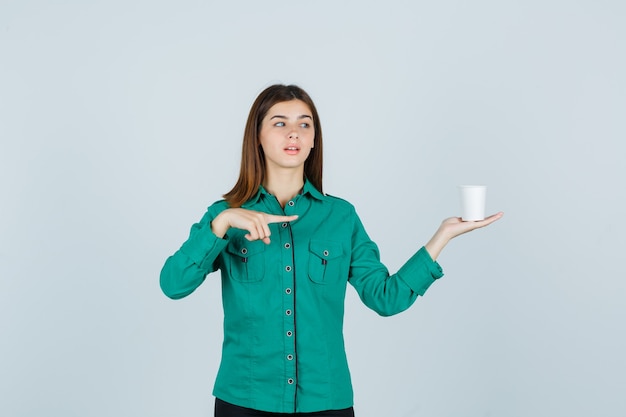 Jonge dame in shirt met plastic kopje koffie terwijl ze naar de rechterkant wijst en gefocust, vooraanzicht kijkt.