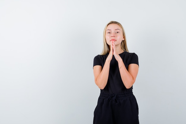 Jonge dame in t-shirt, broek hand in hand in biddend gebaar en op zoek naar hoopvol, vooraanzicht.