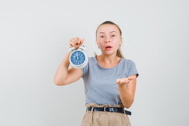 Jonge dame in t-shirt en broek die wekker houdt en verbaasd kijkt