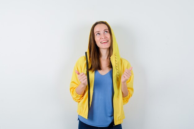 Jonge dame in t-shirt, jas met dubbele duimen omhoog en zelfverzekerd, vooraanzicht.