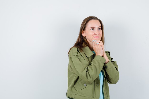 Jonge dame in t-shirt, jas met gevouwen handen op kin en ziet er mooi uit, vooraanzicht.