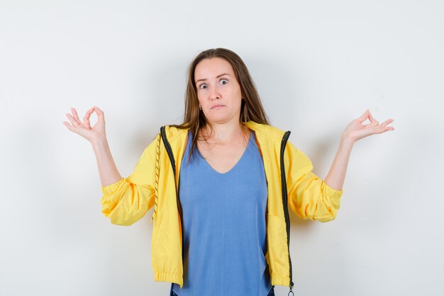 Jonge dame in t-shirt, jas met meditatiegebaar en verbaasd, vooraanzicht.