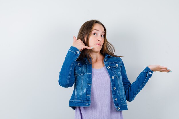 Jonge dame in t-shirt, jas met telefoongebaar, palm opzij spreidend en zelfverzekerd, vooraanzicht.