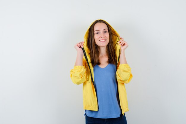 Jonge dame in t-shirt, jas poseren terwijl ze haar capuchon vasthoudt en er zelfverzekerd uitziet, vooraanzicht.