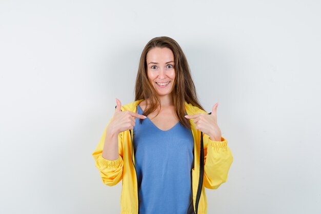 Jonge dame in t-shirt, jas wijst naar zichzelf en ziet er vrolijk uit, vooraanzicht.