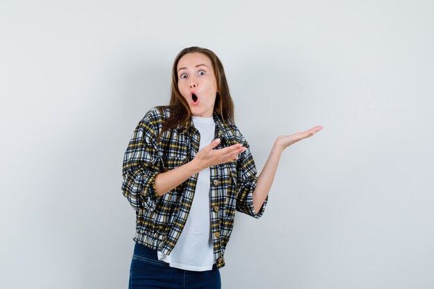 Jonge dame in t-shirt, jasje, spijkerbroek die doet alsof ze iets vasthoudt en verbaasd kijkt, vooraanzicht.