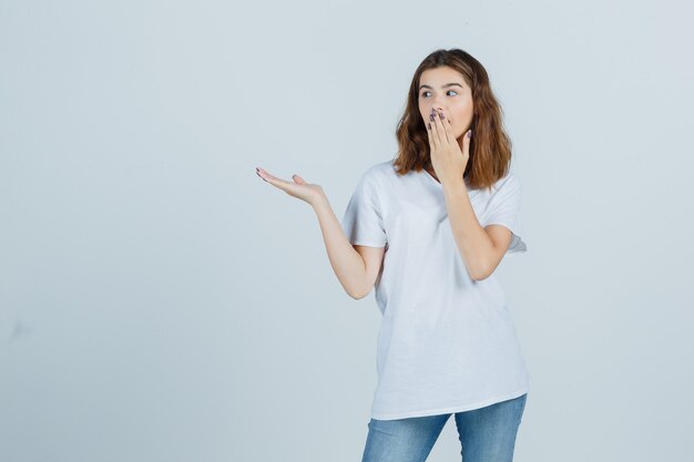 Jonge dame in t-shirt, jeans die hand op mond houdt terwijl iets wordt getoond en verbaasd, vooraanzicht kijkt.