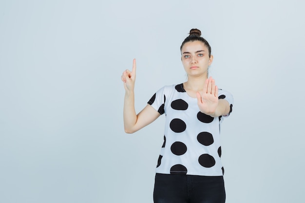 Jonge dame in t-shirt, jeans die omhoog wijst terwijl stopgebaar wordt getoond en ernstig, vooraanzicht kijkt.