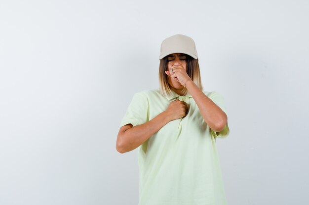 Jonge dame in t-shirt, pet die aan hoest lijdt en onwel kijkt, vooraanzicht.