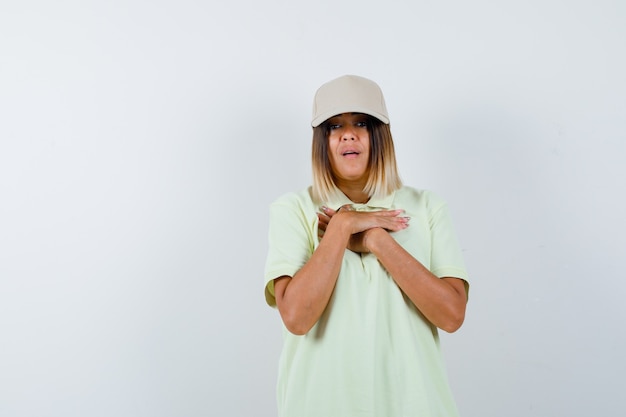 Jonge dame in t-shirt, pet hand in hand op de borst en verbaasd op zoek, vooraanzicht.