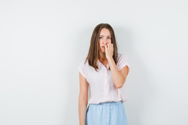 Jonge dame in t-shirt, rok nagels bijten en peinzend, vooraanzicht kijken.