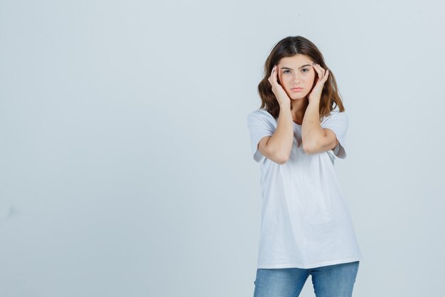 Jonge dame in t-shirt, spijkerbroek die over haar slapen wrijft en er vermoeid uitziet, vooraanzicht.