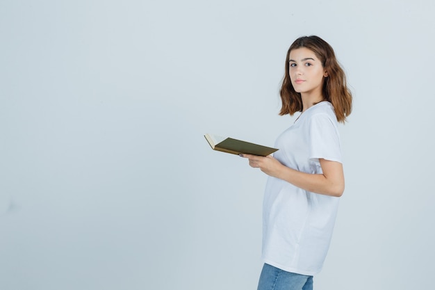 Jonge dame in t-shirt, spijkerbroek met boek terwijl ze over de schouder kijkt en zelfverzekerd kijkt, vooraanzicht.