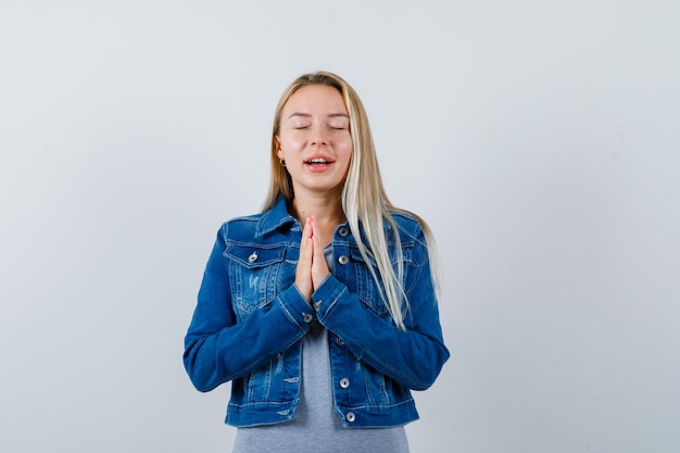 Gratis foto jonge dame in t-shirt, spijkerjasje, rok die de handen tegen elkaar drukt terwijl ze bidt en er hoopvol uitziet