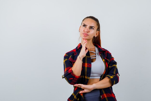 Jonge dame in top, geruit hemd met hand onder kin en peinzend, vooraanzicht.