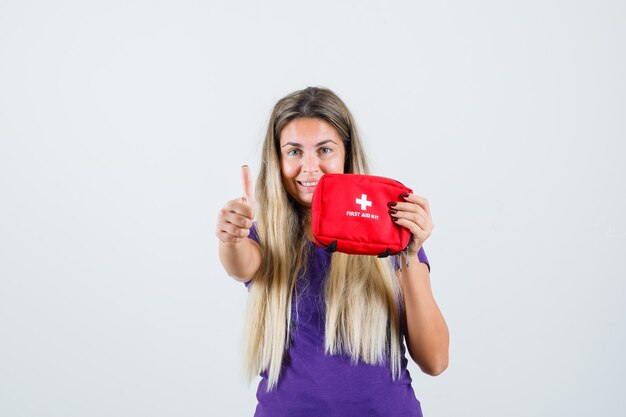 Jonge dame in violet t-shirt met EHBO-kit, duim opdagen en vrolijk, vooraanzicht op zoek.