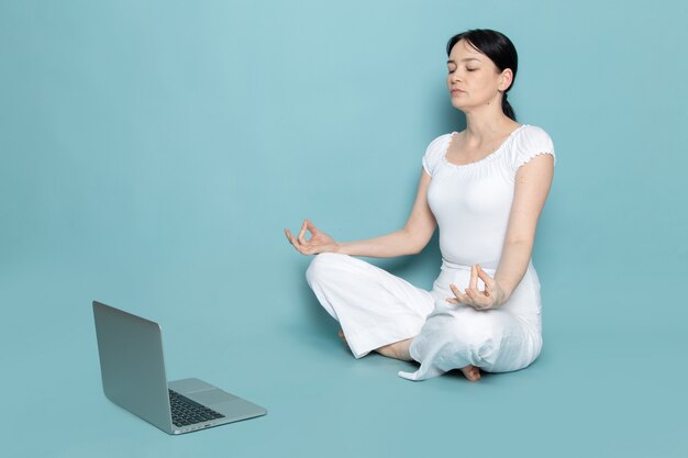 jonge dame in wit overhemd witte broek in youga pose op zoek met behulp van grijze laptop op blauw
