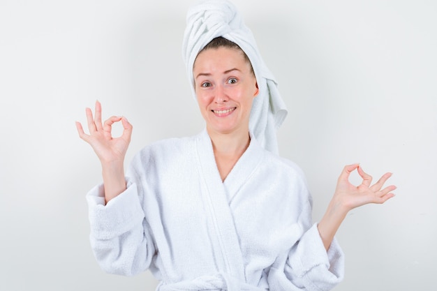 Jonge dame in witte badjas, handdoek die ok gebaar toont en gelukkig, vooraanzicht kijkt.