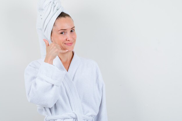 Jonge dame in witte badjas, handdoek die telefoonteken toont en vreedzaam, vooraanzicht kijkt.