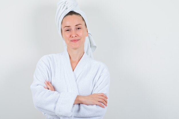 Jonge dame in witte badjas, handdoek die zich met gekruiste armen bevindt en zelfverzekerd kijkt, vooraanzicht.