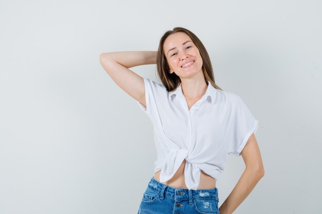 Jonge dame in witte blouse die hand achter hoofd houdt en zelfverzekerd kijkt, vooraanzicht.