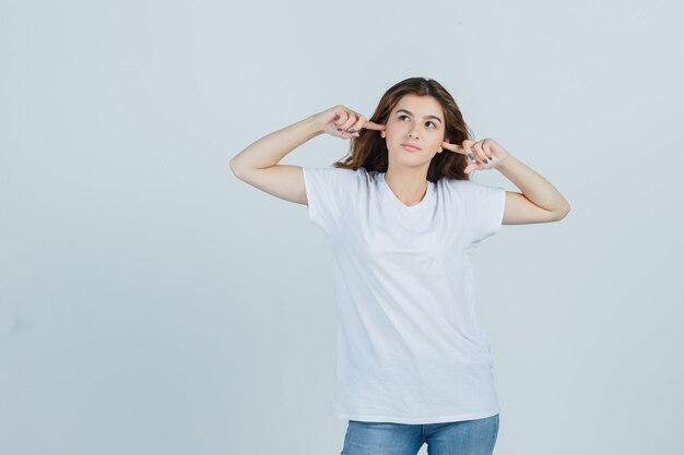 Jonge dame inpluggen oren met vingers in t-shirt, spijkerbroek en op zoek geërgerd, vooraanzicht.