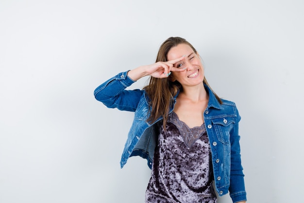 Jonge dame kijkt door vingers in blouse, spijkerjasje en ziet er vrolijk uit, vooraanzicht.