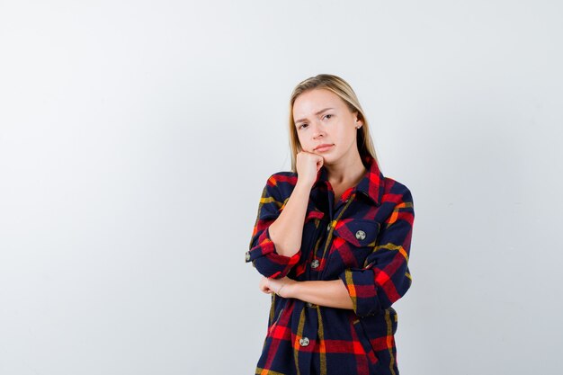 Jonge dame kin leunend op vuist in geruit overhemd en op zoek attent. vooraanzicht.