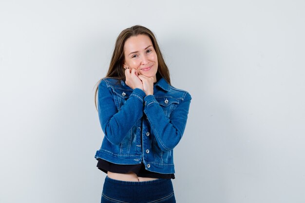 Jonge dame leunend met kin op gevouwen handen in blouse, jas en ziet er schattig uit, vooraanzicht.