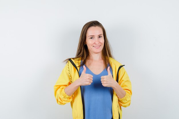 Jonge dame met dubbele duimen omhoog in t-shirt, jas en zelfverzekerd. vooraanzicht.