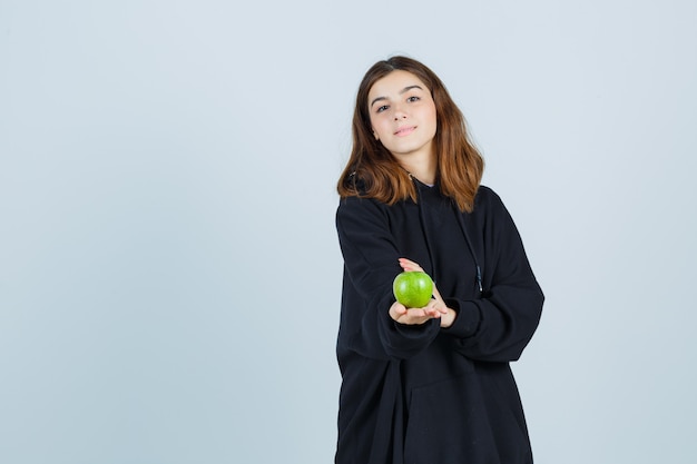 Jonge dame met gebaar terwijl ze appel in oversized hoodie, broek vasthoudt en vreugdevol, vooraanzicht kijkt.