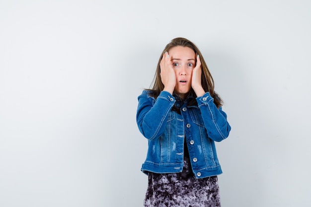 Jonge dame met handen op wangen in blouse, spijkerjasje en verbaasd, vooraanzicht.