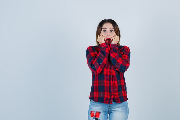 Jonge dame met handen op wangen, mond openend in geruit overhemd, spijkerbroek en met afschuw vervuld, vooraanzicht.