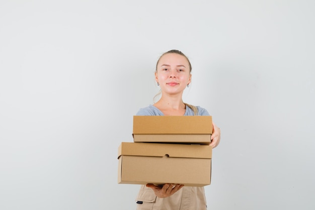 Jonge dame met kartonnen dozen in t-shirt, broek en op zoek vrolijk. vooraanzicht.