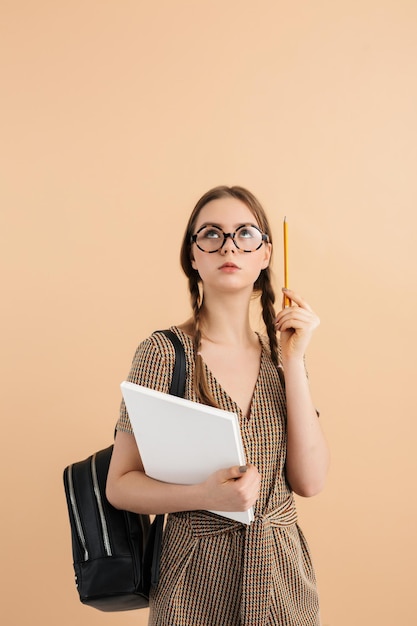 Jonge dame met twee vlechten in tweed jumpsuit en moderne bril met zwarte rugzak op schouder met boek in de hand en potlood in de buurt van hoofd terwijl ze bedachtzaam opkijkt over beige achtergrond