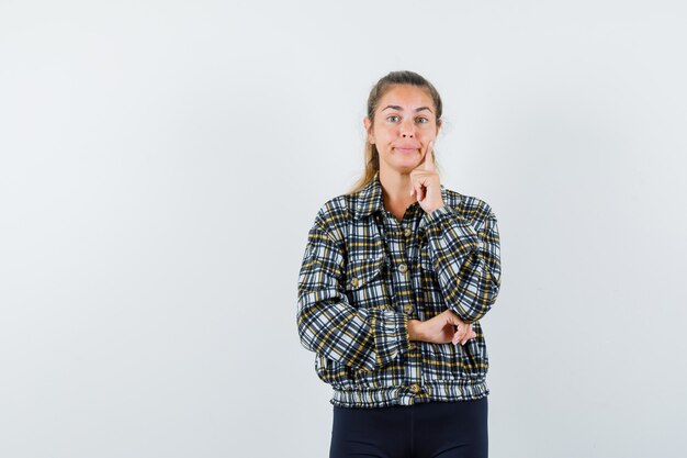 Jonge dame met vinger op de wang in shirt, korte broek en op zoek attent. vooraanzicht.