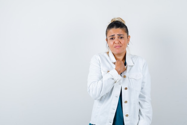 Jonge dame met vuist op borst in shirt, witte jas en verdrietig, vooraanzicht.