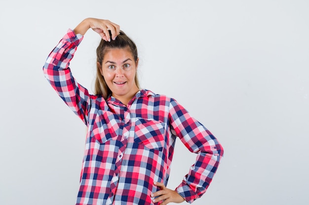 Jonge dame poseren met de hand op het hoofd in geruit overhemd en ziet er grappig uit. vooraanzicht.