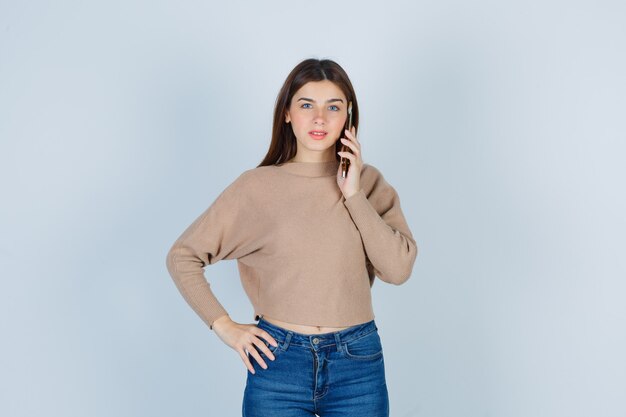 Jonge dame praten op de mobiele telefoon in beige trui, jeans en vrolijk kijken, vooraanzicht.