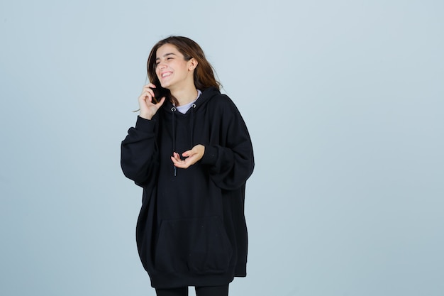 Jonge dame praten op mobiele telefoon in oversized hoodie, broek en op zoek vrolijk. vooraanzicht.