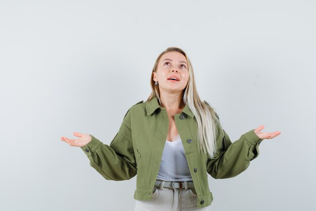 Jonge dame spreidt handen, kijkt omhoog in jasje, broek en kijkt gelukkig, vooraanzicht.