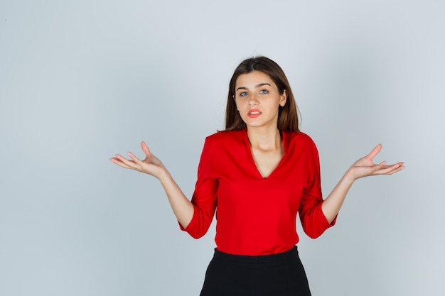 Jonge dame spreidt handpalmen in een clueless gebaar in rode blouse, rok en ziet er hulpeloos uit