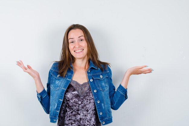 Jonge dame spreidt handpalmen opzij in blouse, spijkerjasje en ziet er gelukkig uit. vooraanzicht.