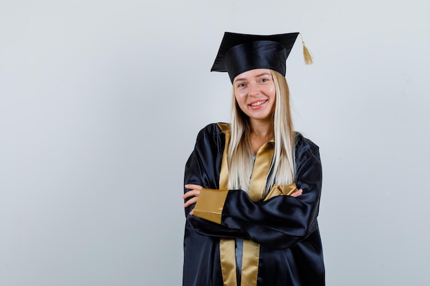 Jonge dame staat met gekruiste armen in academische jurk en ziet er gelukkig uit