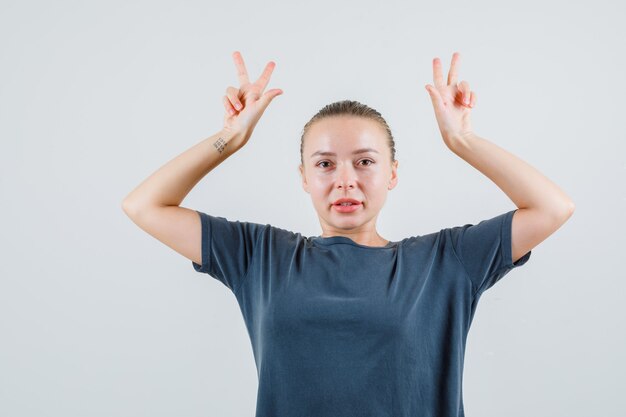 Jonge dame toont v-teken zoals hoorns in grijs t-shirt en ziet er grappig uit