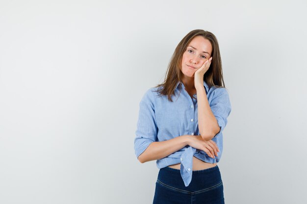 Jonge dame wang leunend op opgeheven handpalm in blauw shirt, broek en peinzend kijken.