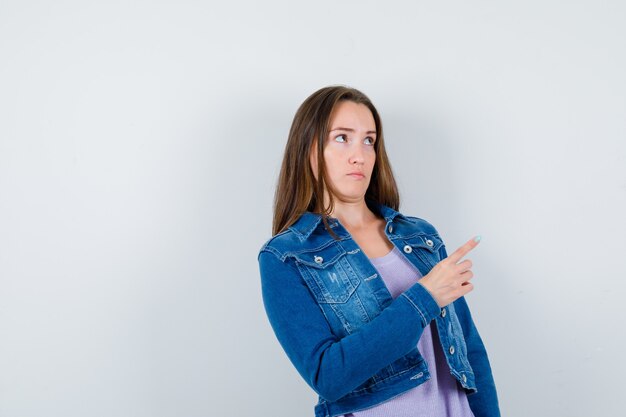 Jonge dame wijst ergens in t-shirt, jas en kijkt gefocust, vooraanzicht.