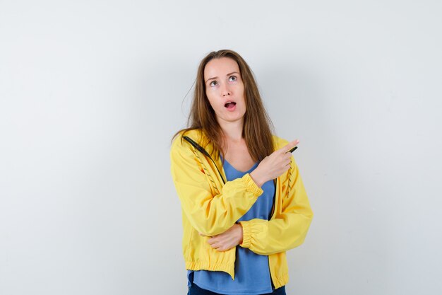 Jonge dame wijst naar de rechterbovenhoek in t-shirt, jas en kijkt peinzend, vooraanzicht.