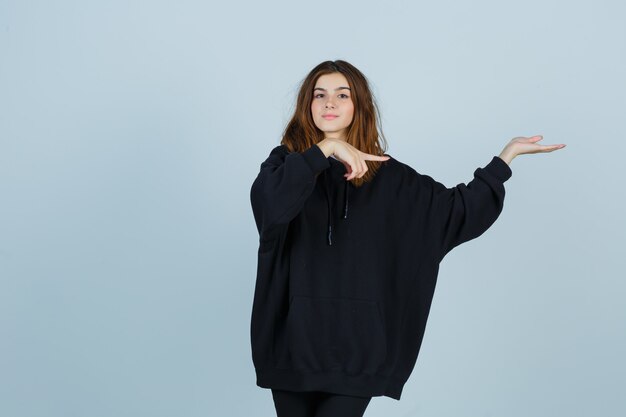 Jonge dame wijst naar de rechterkant terwijl ze iets laat zien in een oversized hoodie, broek en er zelfverzekerd uitziet. vooraanzicht.